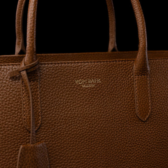 Close-up of a brown pebbled leather tote bag with gold-toned 'VON BAER' branding on the front, set against a solid black background.