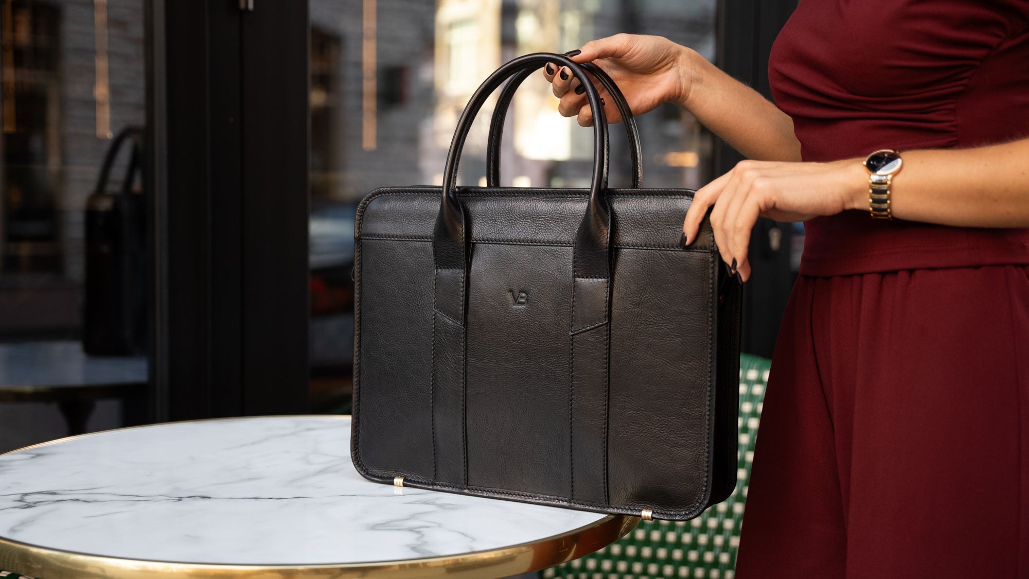 A female model in a red suit is shown with a black Von Baer exquisite luxury leather laptop bag for women, which is placed on a marble table.