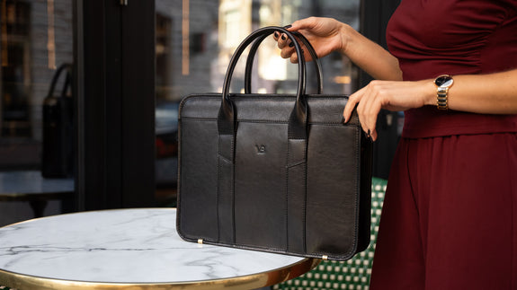 A female model in a red suit is shown with a black Von Baer exquisite luxury leather laptop bag for women, which is placed on a marble table.
