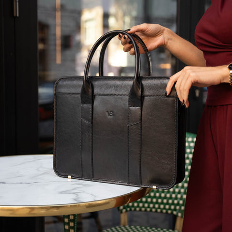 A female model in a red suit is shown with a black Von Baer exquisite luxury leather laptop bag for women, which is placed on a marble table.