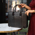 A female model in a red suit is shown with a black Von Baer exquisite luxury leather laptop bag for women, which is placed on a marble table.