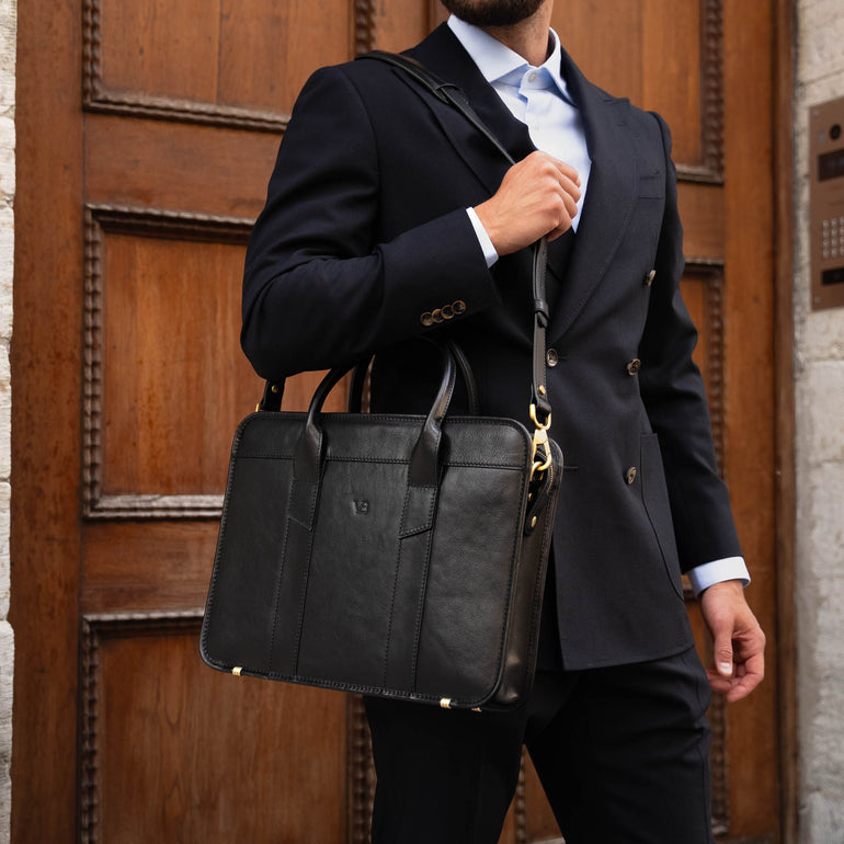 An exquisite, slim, black Von Baer full-grain leather laptop briefcase with a shoulder strap, held by a man in a suit on a strap in town