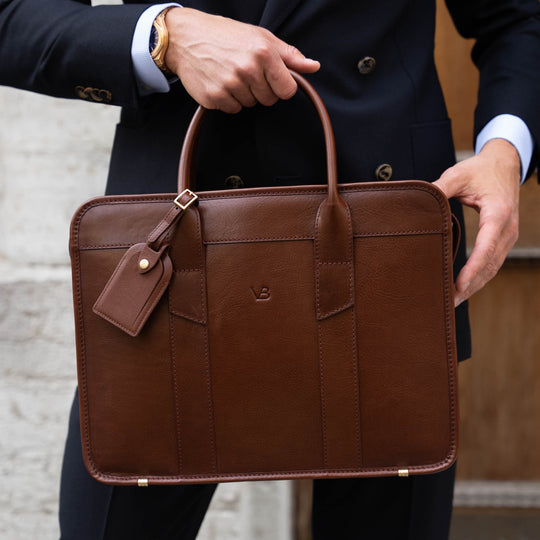 An exquisite, slim, brown Von Baer full-grain leather laptop briefcase with a luggage tag, held by a businessman