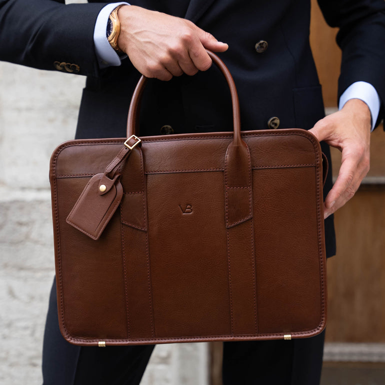An exquisite, slim, brown Von Baer full-grain leather laptop briefcase with a luggage tag, held by a businessman
