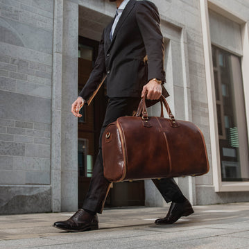 A person in a suit walking and carrying a brown leather garment duffle bag over their shoulder.