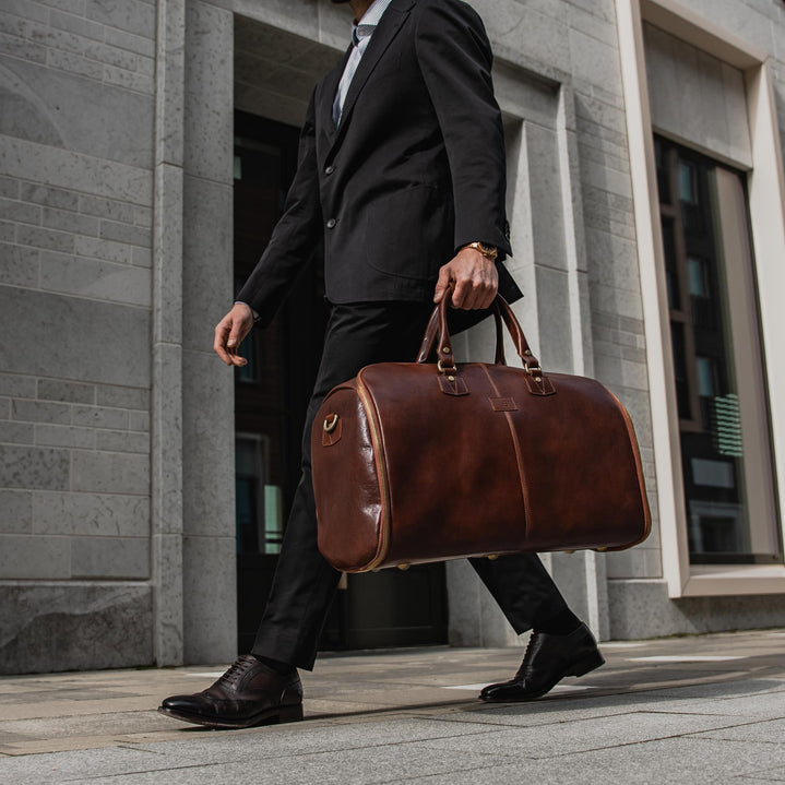 A person in a suit walking and carrying a brown leather garment duffle bag over their shoulder.