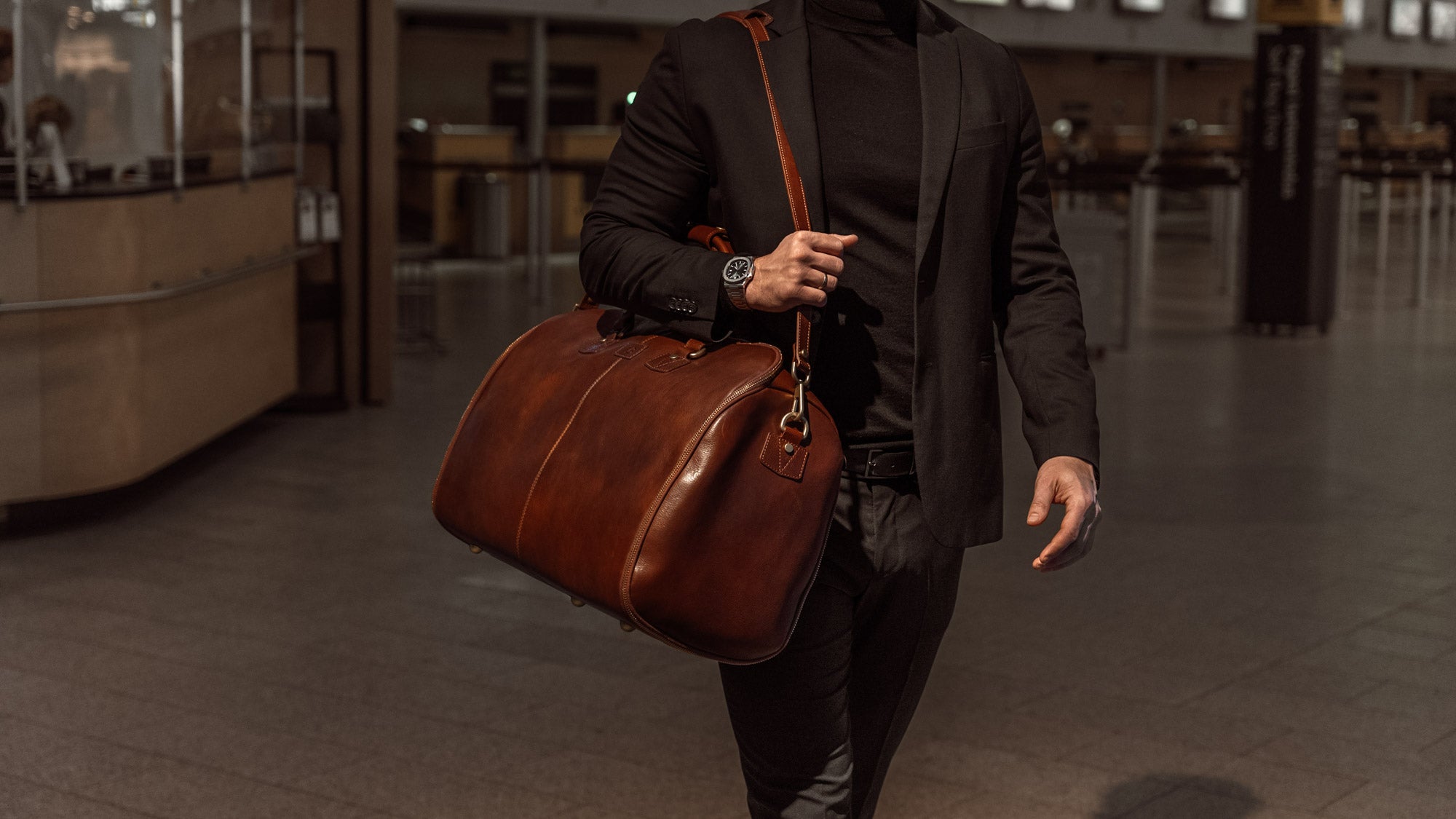 A male model in an airport is carrying a brown Von Baer large luxury leather garment bag with a shoulder strap.