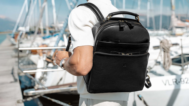 A man wearing a white shirt and light-colored shorts carries a black full grain leather backpack on his back while standing at a marina with boats and mountains in the background.