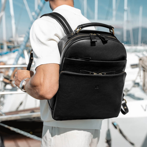 A man wearing a white shirt and light trousers is shown from behind, carrying a black full grain leather backpack on his back in a marina setting with boats and masts in the background.