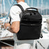 A man wearing a white shirt and light trousers is shown from behind, carrying a black full grain leather backpack on his back in a marina setting with boats and masts in the background.