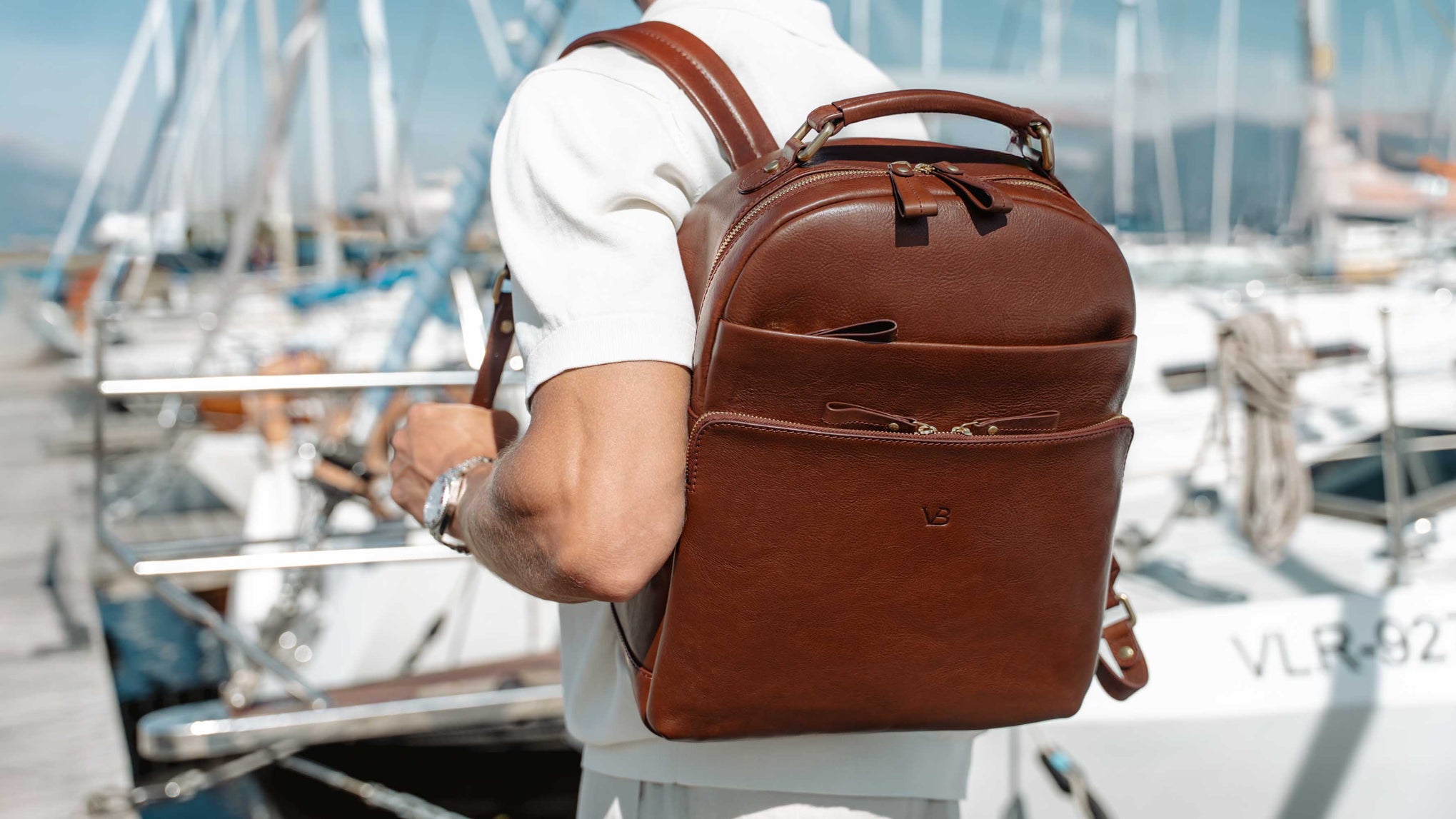 A man wearing a white shirt and light-colored shorts carries a solid brown full grain leather backpack on his back while standing at a marina with boats and mountains in the background.