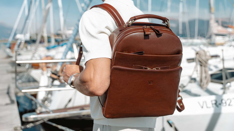 A man wearing a white shirt and light-colored shorts carries a solid brown full grain leather backpack on his back while standing at a marina with boats and mountains in the background.