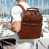 A man wearing a white shirt and light trousers is shown from behind, carrying a solid brown full grain leather backpack on his back in a marina setting with boats and masts in the background.
