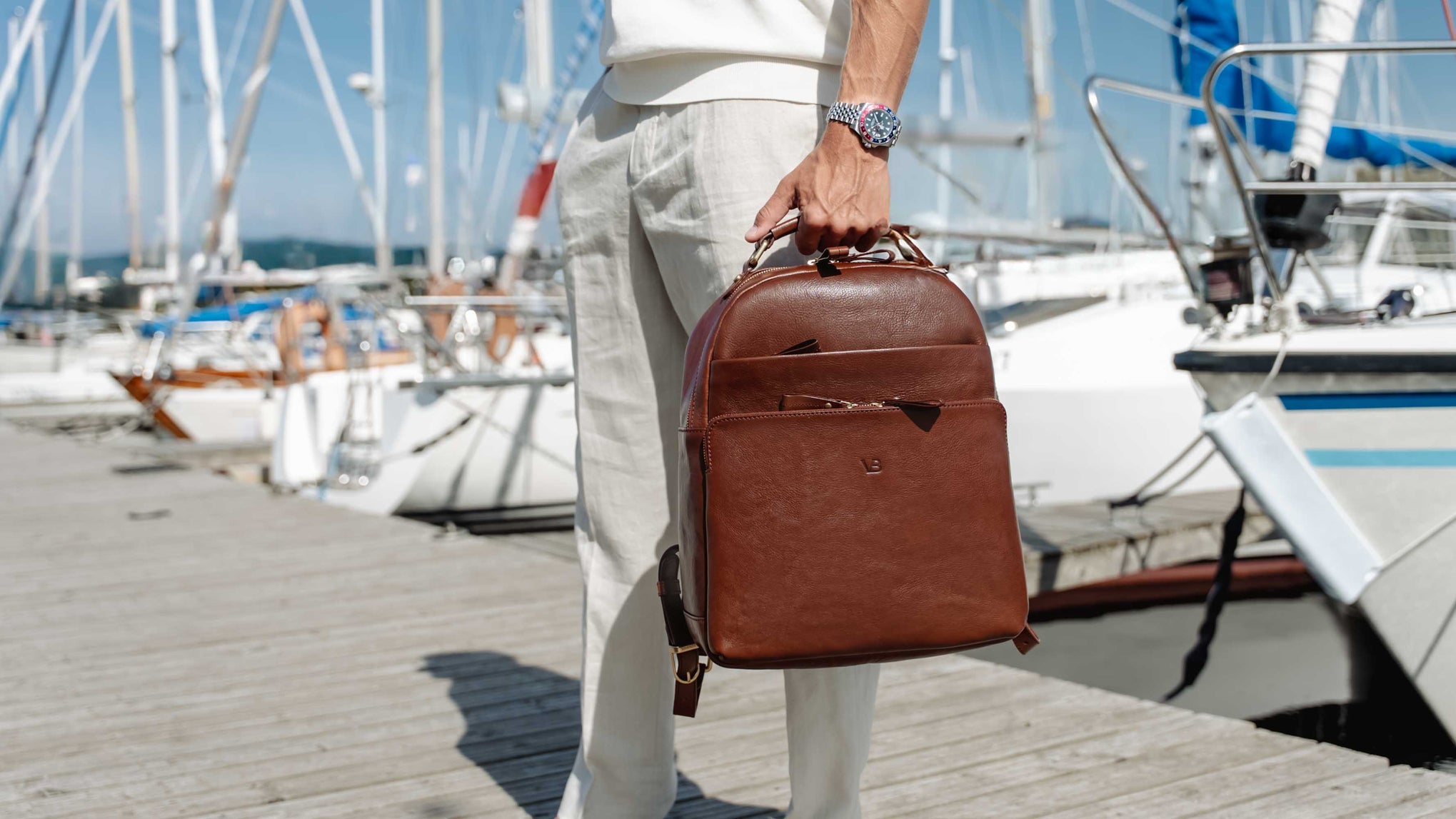 A person standing on a wooden dock by a marina holds a solid brown full grain leather backpack for men by von baer. The background shows white boats and masts in the water under a bright sky.