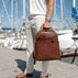 A man in a white shirt and light trousers and rolex watches on the hand holds a solid brown full grain leather backpack by von baer on a wooden dock with boats in the background.
