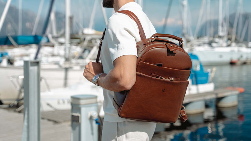 A man wearing a white shirt and white pants in quiet luxury style carries a brown full grain leather backpack with a top handle and front zip pocket, standing at a marina with boats and water in the background.