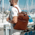 A man in a white shirt and sunglasses carries a solid brown full grain leather backpack by von baer with a top handle and front zip pocket, standing at a marina with boats in the background.