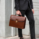 Person in a suit holding a brown leather briefcase against a light stone wall.
