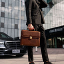 Person in a suit holding a brown leather briefcase in front of a modern building with a car in the background.