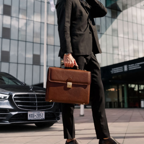 Person in a suit holding a brown leather briefcase in front of a modern building with a car in the background.