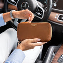 A person holding a brown luxurious pebbled leather zip-around wallet in a car interior. The wallet has gold-tone hardware and 'Von Baer' printed in gold on the front.