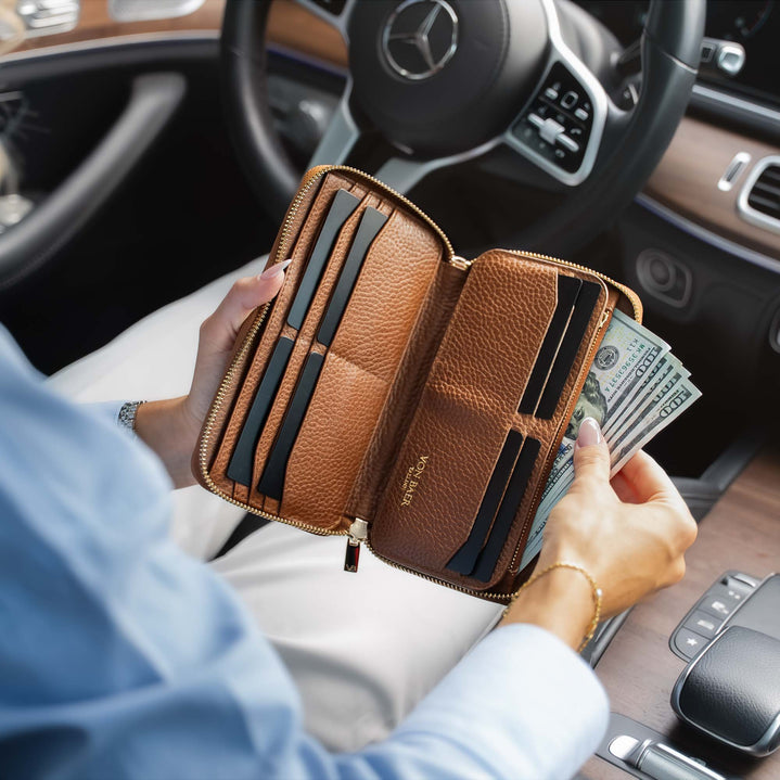 A person sitting in a expensive car holds an open luxurious classic tan leather zip-around wallet showing card slots, cash, and interior compartments. The Mercedes-Benz logo is visible on the steering wheel.