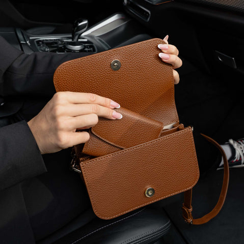 Von Baer Reverie compact cognac tan full-grain leather wallet held open inside a car interior alongside an Isabella small leather crossbody bag for women, shown by a woman wearing a black blazer.