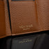 Von Baer Reverie compact cognac pebbled leather wallet for women, shown in close-up highlighting the gold foil Von Baer Tallinn logo embossed on a leather tab and the mirrored branding on the black leather interior section
