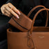 Von Baer Reverie compact women's pebbled leather wallet in cognac, being placed into an Estelle small leather tote bag with double top handles and a gold-tone logo plaque, shown in a close-up against a dark background