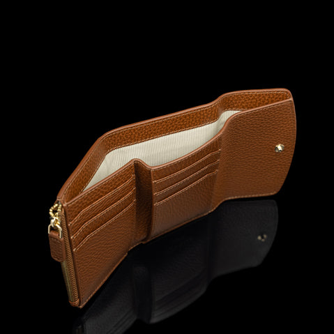 Von Baer Reverie small cognac pebbled leather wallet for women, shown open to reveal card slots, a cream-colored lining, and a banknote compartment with a gold-tone zipper pull against a reflective black background