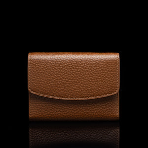 Von Baer Reverie small cognac tan pebbled leather wallet for women with a curved flap closure, shown centered in a front view against a solid black background with a subtle reflection.