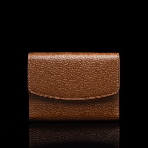 Von Baer Reverie small cognac tan pebbled leather wallet for women with a curved flap closure, shown centered in a front view against a solid black background with a subtle reflection.