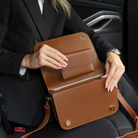 Von Baer Reverie small cognac pebbled leather wallet and Isabella Grande women's leather crossbody bag, held open by a woman in a black blazer seated in a car, showcasing the spacious interior compartments and leather strap