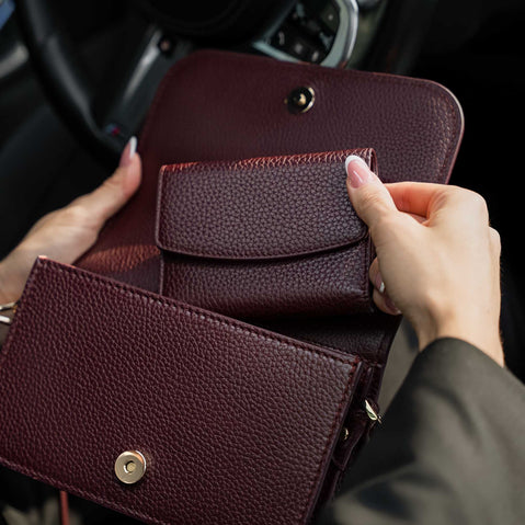 Von Baer Reverie burgundy pebbled compact leather women's wallet and Isabella small leather crossbody bag, shown in close-up inside a car, highlighting the open wallet's interior, snap-button closure, and small zippered pocket
