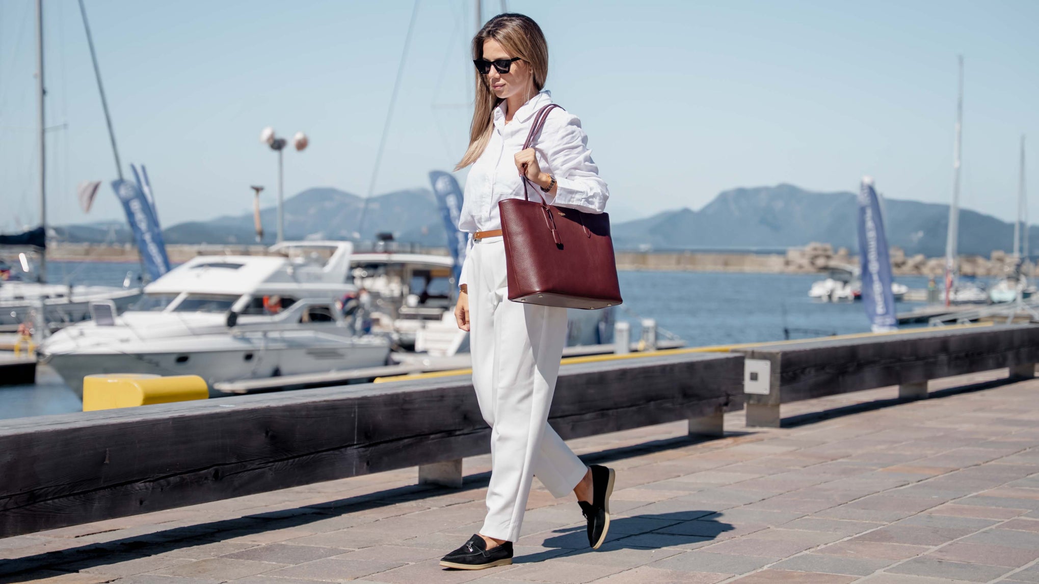 A woman walking along a sunny harbor promenade, wearing a white button-up shirt and white trousers, carrying a luxury large dark red leather tote bag by Von Baer.