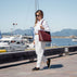 A beautiful woman walking along a sunny harbor promenade, wearing a white shirt and white trousers in quiet luxury style and carrying a large burgundy tote bag for laptop.