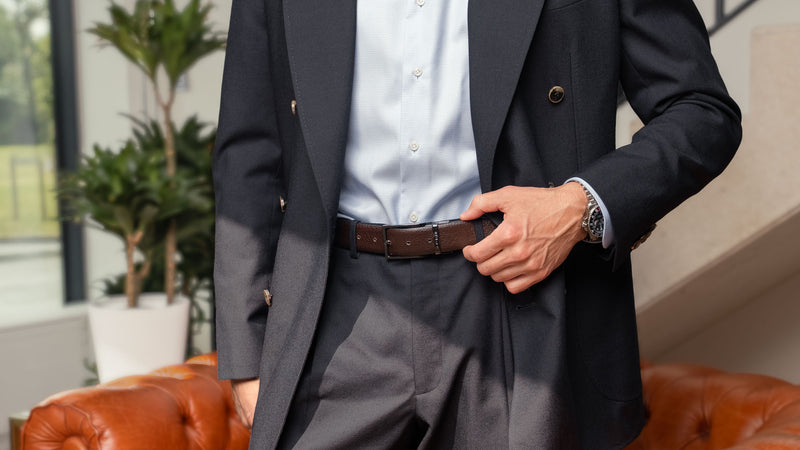 Von Baer Premier full-grain reversible brown leather 2-in-1 belt, worn by a man in a dark suit with a light shirt, standing indoors in a luxury apartment setting
