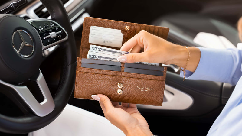 A brown pebbled luxurious tan leather wallet by Von Baer, held open by a person inside a car. The wallet contains credit cards and cash, with the Mercedes-Benz steering wheel visible in the background.
