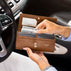 A big tan pebbled leather purse with gold-tone hardware, shown open in a car interior. A hand is sliding cash into the card slots, and the brand name 'VON BAER' is visible on the front.