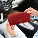 A person holding a red pebbled leather purse with 'VON BAER' printed on the front, set against a blurred car interior background.