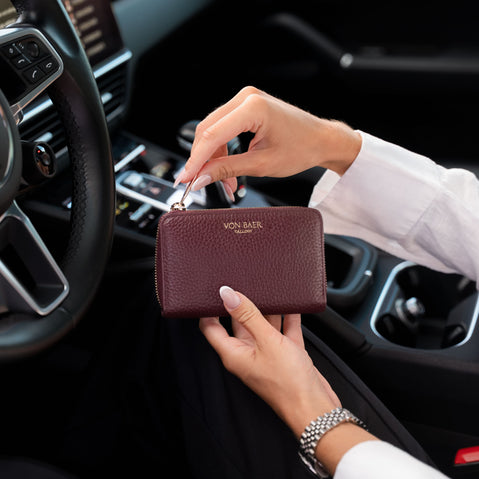 A person in a white dress shirt holds a high quality burgundy pebbled leather wallet with gold hardware inside a black car interior. The wallet features the gold embossed brand name 'VON BAER'.