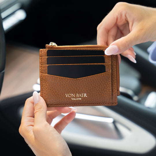 A brown pebbled leather card holder wallet with gold-toned hardware, held by two hands in front of a blurred car interior. The wallet features the brand name 'VON BAER' and 'TALLINN' printed in gold on the front pocket.