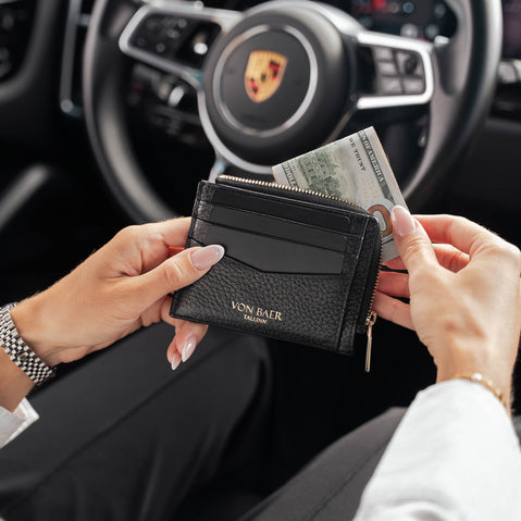 A person holds a black textured rich leather wallet with gold lettering reading 'VON BAER' in a car interior, with a Porsche steering wheel blurred in the background.