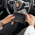 A person holds a black textured rich leather wallet with gold lettering reading 'VON BAER' in a car interior, with a Porsche steering wheel blurred in the background.