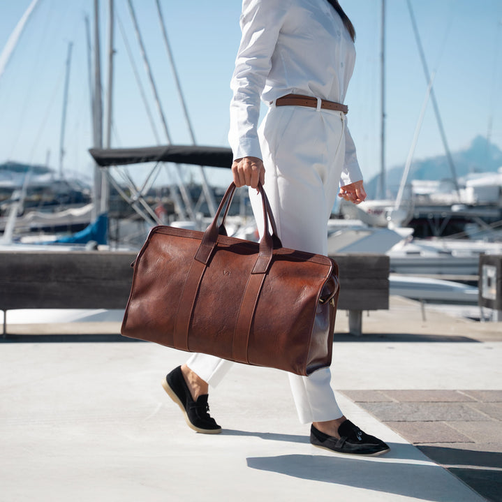 A woman in a white outfit carrying a von baer brown leather duffel bag while walking along a sunny dockside path with boats, mountains and masts in the background.